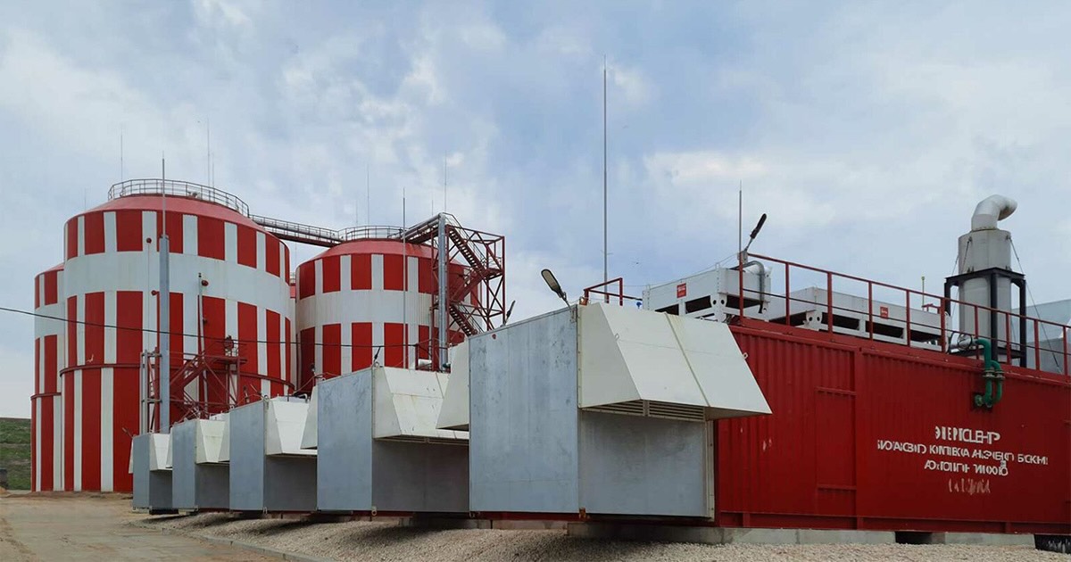 Container CHP plant at Timokhovo biogas landfill in Russia with five MWM TCG 2020 V20 engines. Red containers with white lettering stand outdoors, with red-and-white striped industrial structures in the background under a partly cloudy blue sky.
