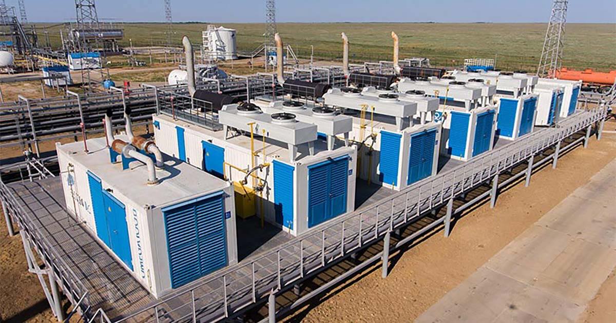 Container CHP plant with four MWM TCG 2020 V12 engines at Bykovogaz oil and gas facility in Volgograd Region, Russia. Several blue-and-white containers stand in parallel on a raised metal platform above sandy ground, with chimneys, pipes, and other industrial structures in the background. Beyond the fences are grassy fields under a sunny blue sky.