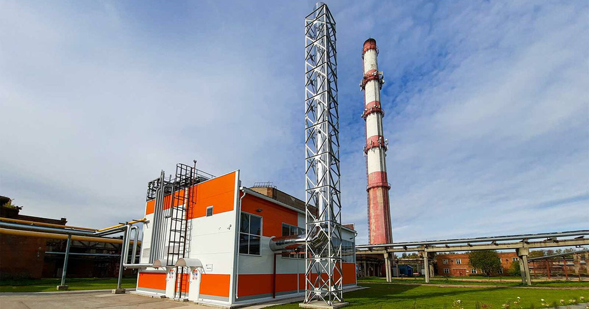 Exterior view of the striped red-orange and white building housing three MWM TCG 2032 V16 gensets at Sukhonsky Cardboard and Paper Mill LLC, Russia; set on a concrete surface surrounded by grass, pipes, and other industrial structures, with a metal chimney attached and a larger red-orange and white-striped smokestack in the background under a blue sky with wispy clouds.