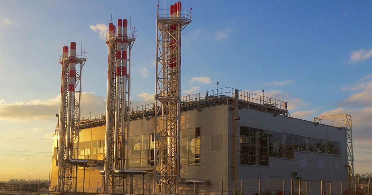 Industrial building at Industrial Park-1, Russia, in golden sunlight. White-gray rectangular structure with metal scaffolding. Three chimneys on the left side have red-and-white striped stacks. Facility uses four TCG 2020K V16 and two TCG 2032 V16 engines. Blue sky with scattered clouds and distant trees in the background.