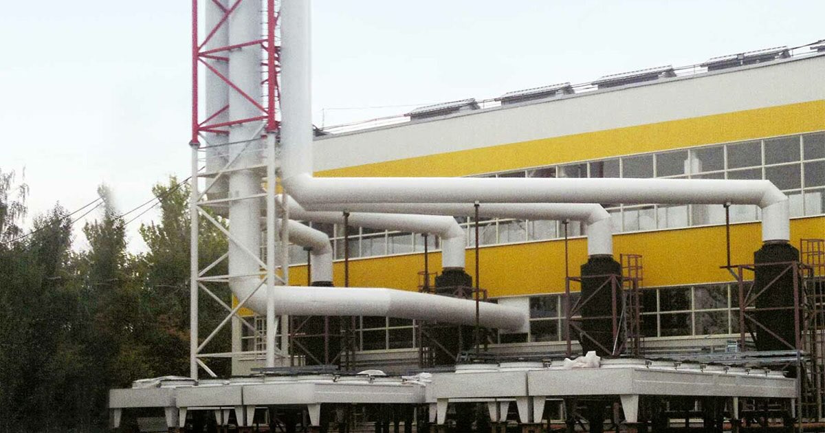 Exterior view of the Energy Security facility for NICEVT in Moscow, Russia, featuring four TCG 2032 V16 engines. The yellow-and-white striped building has five white pipes leading into a chimney-like structure, surrounded by industrial structures. Trees on the left are green under a hazy daytime sky.