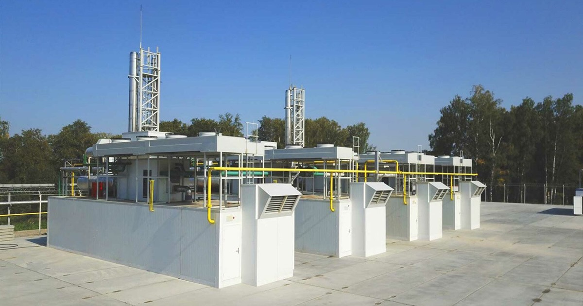 Container CHP plant at Bereznyakovskiy Mining and Processing Plant with four MWM TCG 2020 V20 engines. Three white containers stand on a concrete outdoor area with industrial structures like vents and pipes. The site is fenced, surrounded by green trees under a sunny, clear blue sky.