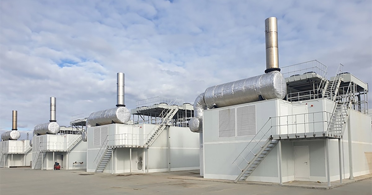 Four white-grey containerized MWM TCG 2032B V16 gas engines on the asphalted grounds of Svetlinskaya Gold Processing Plant in Plast, Russia; cloudy sky with patches of blue visible in the background.