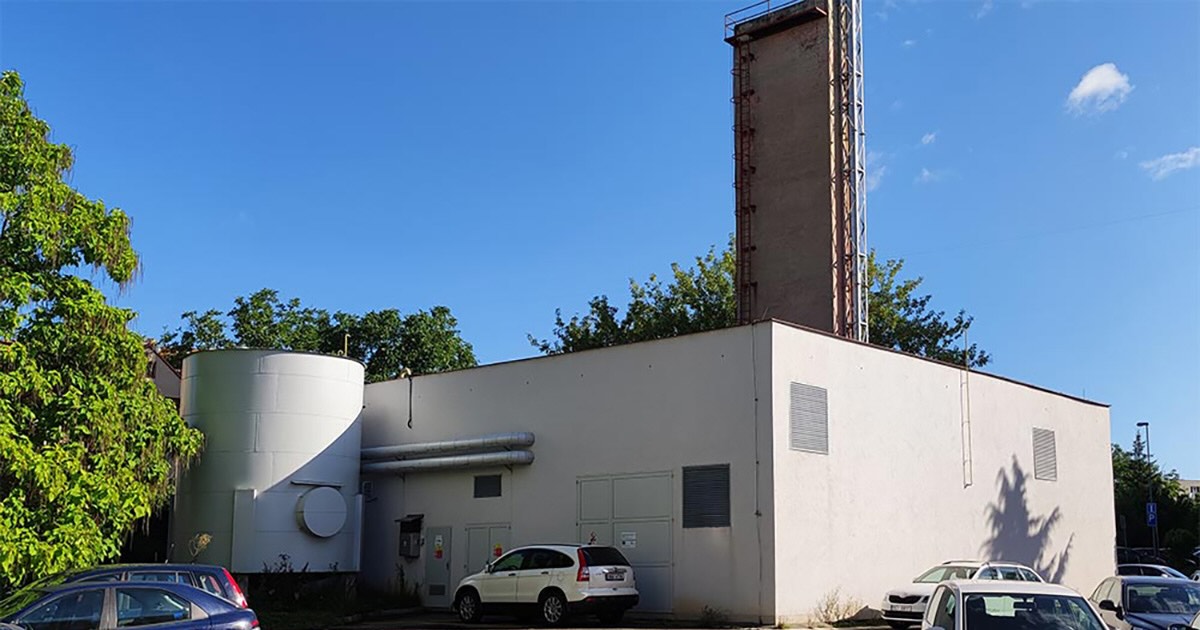Exterior of the Brandýsko power plant in Brandýs nad Labem, Czech Republic, with one MWM TCG 3016 V12 gas engine for reliable heat and power. White industrial building with chimney-like tower, metal pipes along the façade, cars parked in front, and green trees under a sunny blue sky.