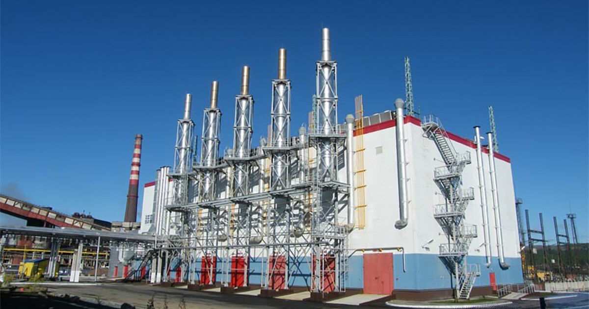 Exterior view of the industrial building housing five MWM TCG 2032 V16 gensets at Sredneuralsky Copper Smelter, Russia; white facade with a wide blue stripe along the lower part of the building reaching up to the top of the red double doors, and a narrow red stripe along the roofline. Five metal chimneys are mounted directly on the building, with a metal staircase on the right side, all surrounded by additional industrial structures.