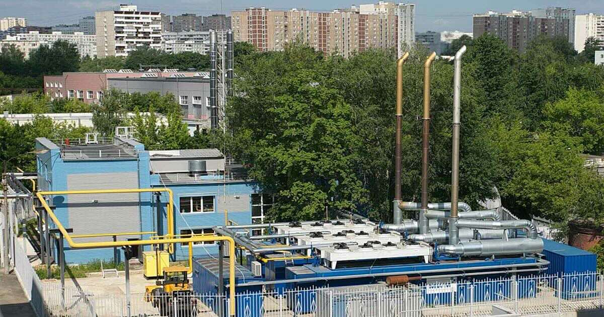 Three containerized MWM TCG 2020 V16 gas engines in signature blue on the fenced grounds of Vertikal JSC warehouse complex in Moscow, Russia, viewed from an elevated angle; light blue building, green trees, and city high-rises in the background.