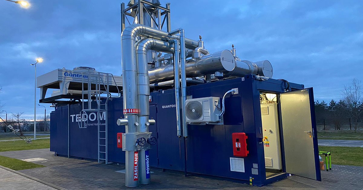 Containerized MWM TCG 3016 V16 gas engine in a dark blue container on a paved area at Szydłowiec district heating plant, Poland; behind it, alternating grass and paved sections, a chain-link fence, and bare trees. The dim, cloudy sky and illuminated streetlights suggest a winter morning or evening.